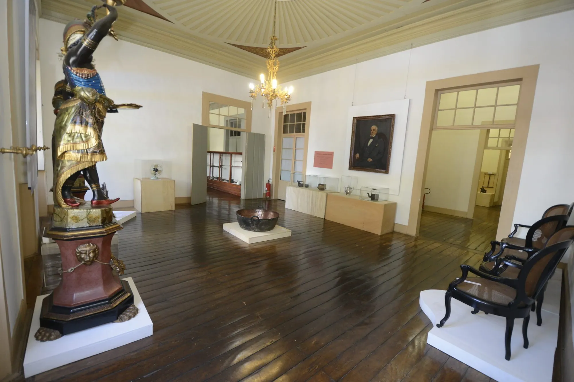 Salão expositivo do Solar do Barão com escultura, lustre e piso de tábuas